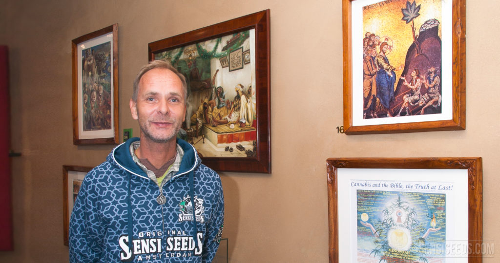 Alan Dronkers portant une hotte de sweats de Sensi Seeds contre un mur couvert de peintures encadrées