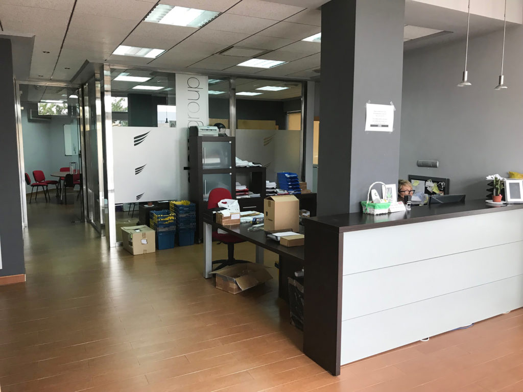 Foto de las oficinas de Lamarihuana.com. Una mujer con gafas se sienta detrás de su escritorio. Las paredes son de color gris, y el suelo es de madera marrón. Hay cajas de entrega marrones en el espacio.