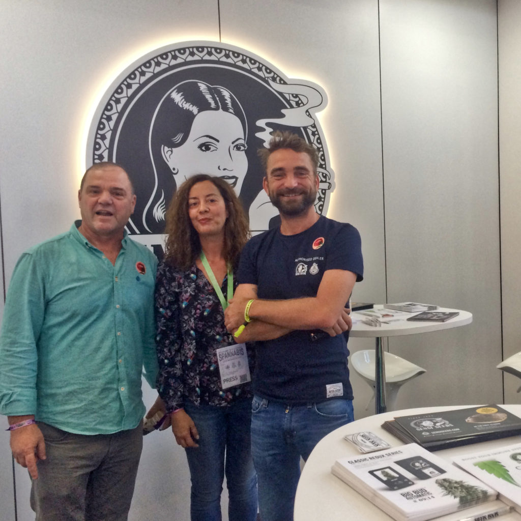 Miranda y Ravi Spaarenberg, de Sensi Seeds, posan juntos con Jesús Mac, de lamarijuana.com, en el stand de Sensi Seeds en Spannabis Madrid 2017. Detrás de ellos, hay un gran logotipo de Sensi Seeds. Delante de ellos, hay una mesa circular blanca con literatura, folletos y pegatinas de Sensi Seeds.