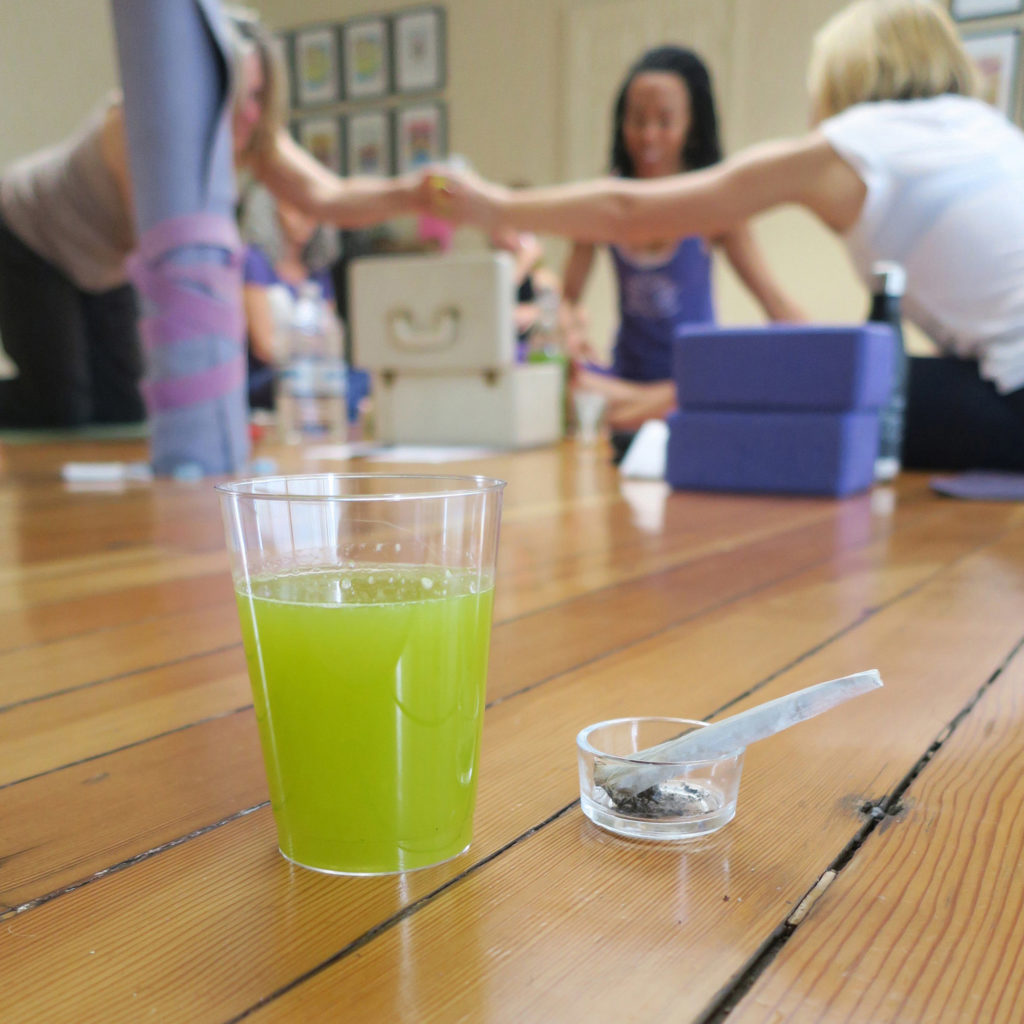 Foto de una mesa que tiene encima un vaso de plástico transparente con zumo de cannabis verde y brillante, y un porro de cannabis en un pequeño cenicero, y al fondo, un grupo de personas que comparten zumo de cannabis.