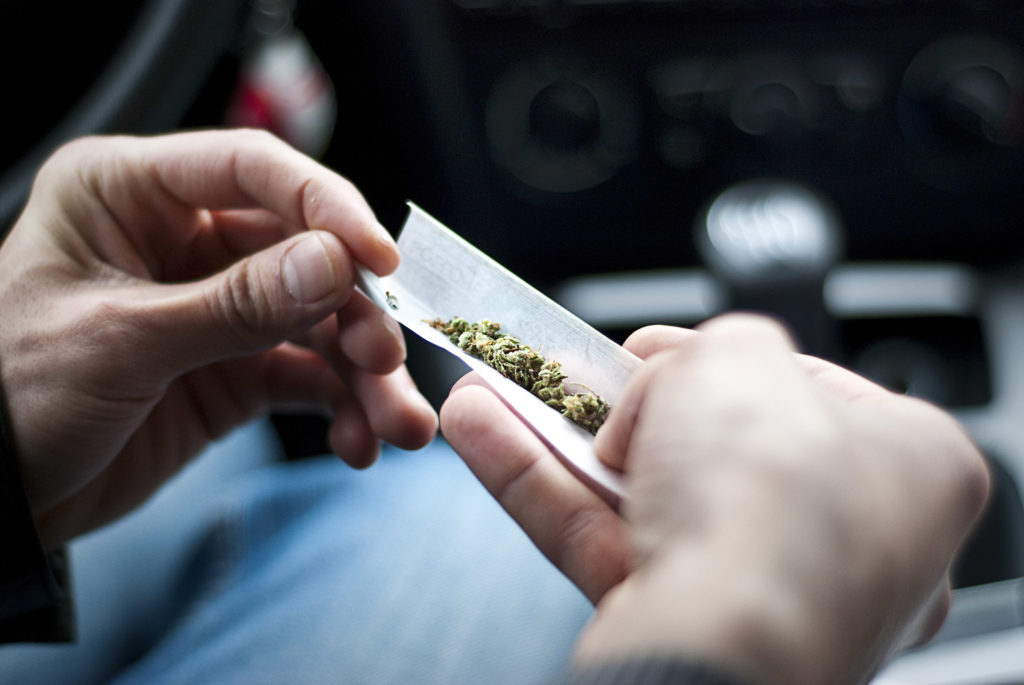 Fotografía que muestra un primer plano de dos manos liando un porro, dentro de un automóvil.