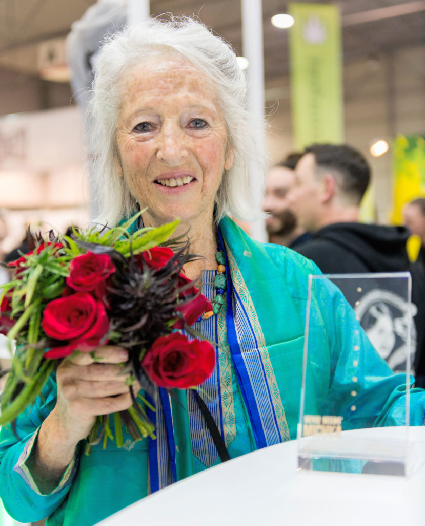 Ein Foto der Cannabis-Ikone Michka. Sie hält einen Blumenstrauß und trägt eine blaugrüne Jacke. Vor ihr steht ein Glaspreis auf einem Tisch. Sie ist auf dem Cannafest 2017. Hinter ihr ist ein Mann zu sehen, der einen Hoodie von Sensi Seeds trägt.