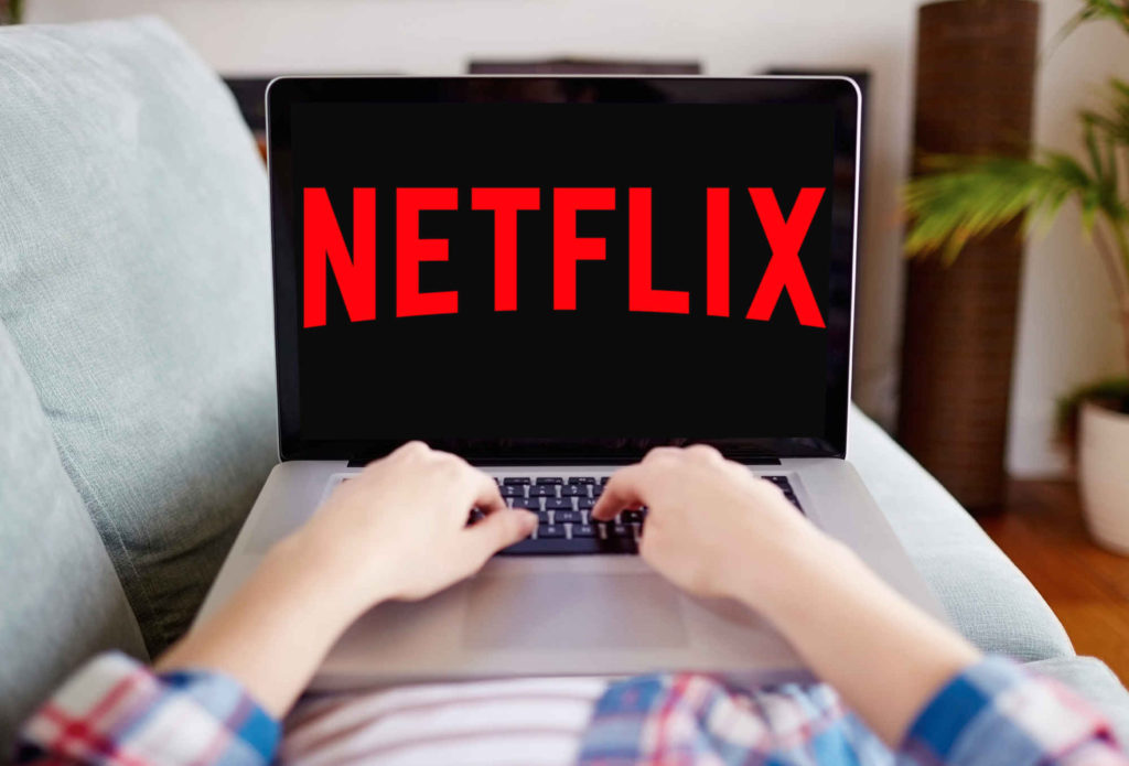 A photograph of a laptop computer resting on someone’s lap, as they lie on a couch. The screen shows the Netflix logo.