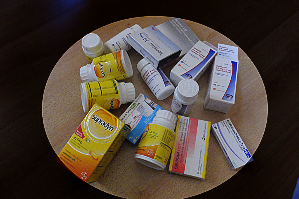 Foto von Medikamentenverpackungen und -dosen, die auf einem runden Holztisch liegen. Insgesamt handelt es sich um je 8 Verpackungen und Dosen. Erkennbar ist Supradyn activo, Xeristar 30 mg, Sevikar HCT und Ácido ibandrónico Teva 150 mg.