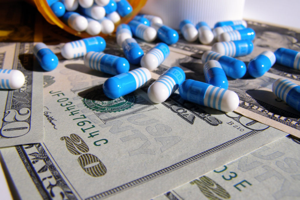 Foto van blauwwit gestreepte capsules afkomstig uit een container op de achtergrond. De capsules liggen op bankbiljetten van 20 dollar.