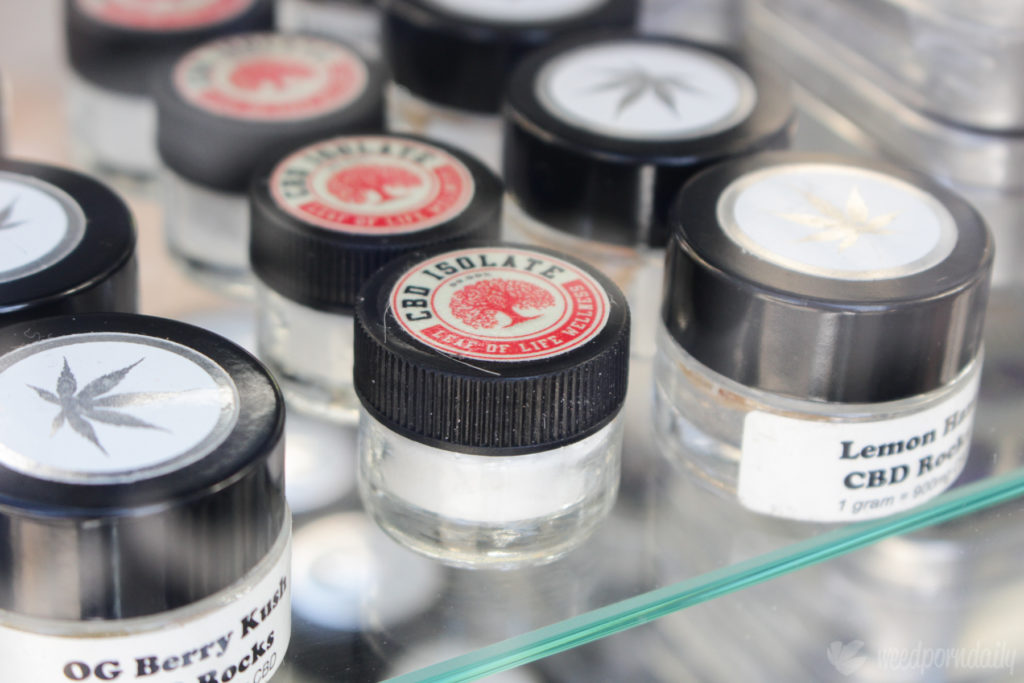 Close-up van kleine doosjes met CBD-kristallen. Links staat “OG Berry Kush“ en in de dozen rechts zitten kristallen van de soort “Lemon Haze“. We kunnen niet zien welke kristallen in de middelste dozen zitten. Op deze dozen staat “CBD Isolate – Leaf of Life Wellness“.