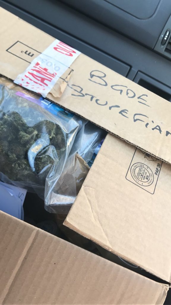 A picture of a cardboard box sitting in the passenger seat of a car. The box is half-closed and shows plastic bags full of dried cannabis buds. Written in black marker on the box: BGDE STUPEFIANT