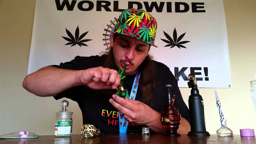 A man with long brown hair and a cap lighting a cannabis pipe