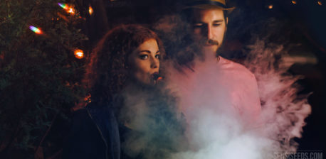 Fotografía de un hombre y una mujer jóvenes, tomada de noche. La mujer inhala de un vaporizador que produce una gran humareda.