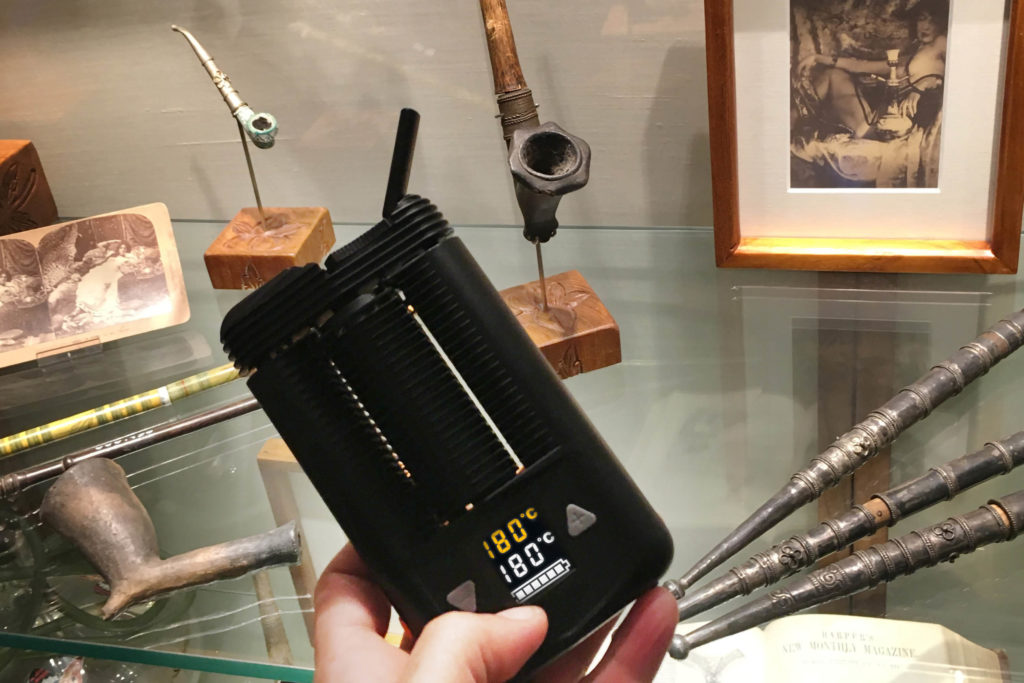 A hand holding up a cannabis vaporiser. There are different types of pipes in the background