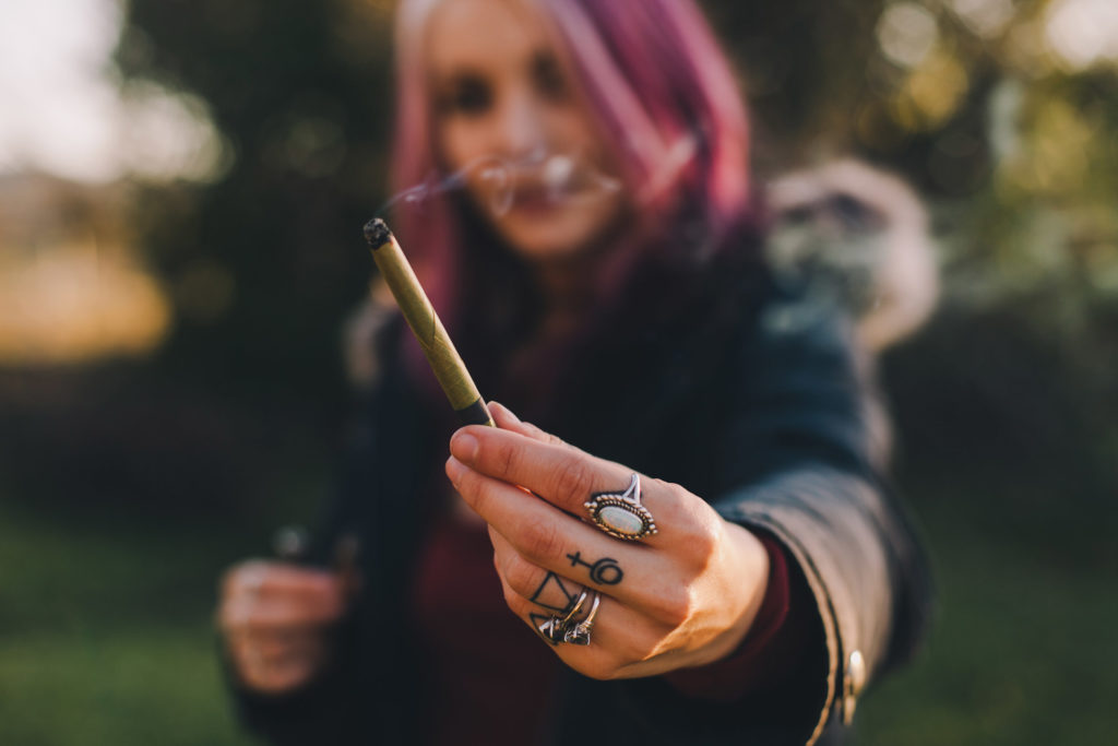 Woman with pink colored hair, reaching her hand with rings and tattoos, holding a joint