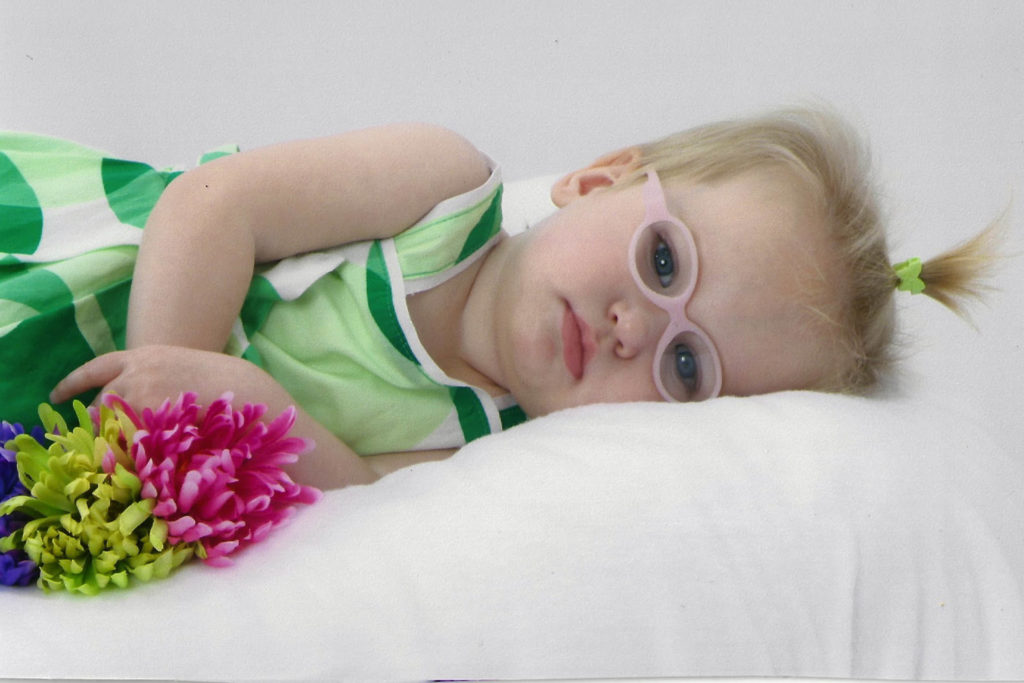 A toddler wearing pink framed glasses resting on a white bed and holding flowers