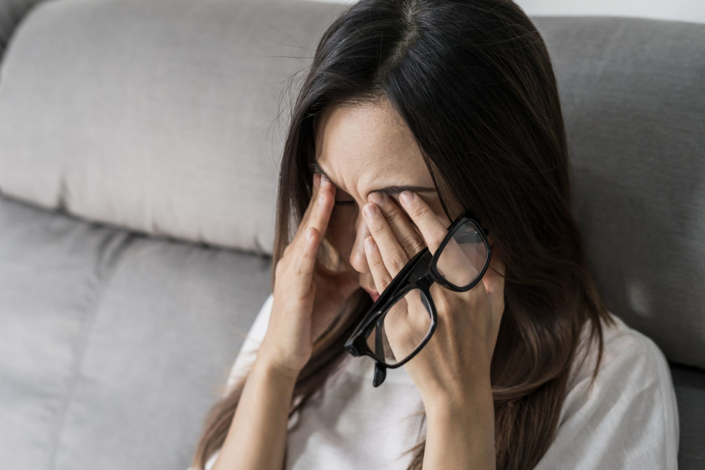 Una mujer con cabello largo y castaño presionando su copa con dolor y sosteniendo sus gafas.