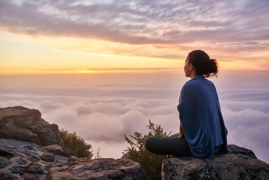 A woman sitting on the top of the mountain with clouds beneath