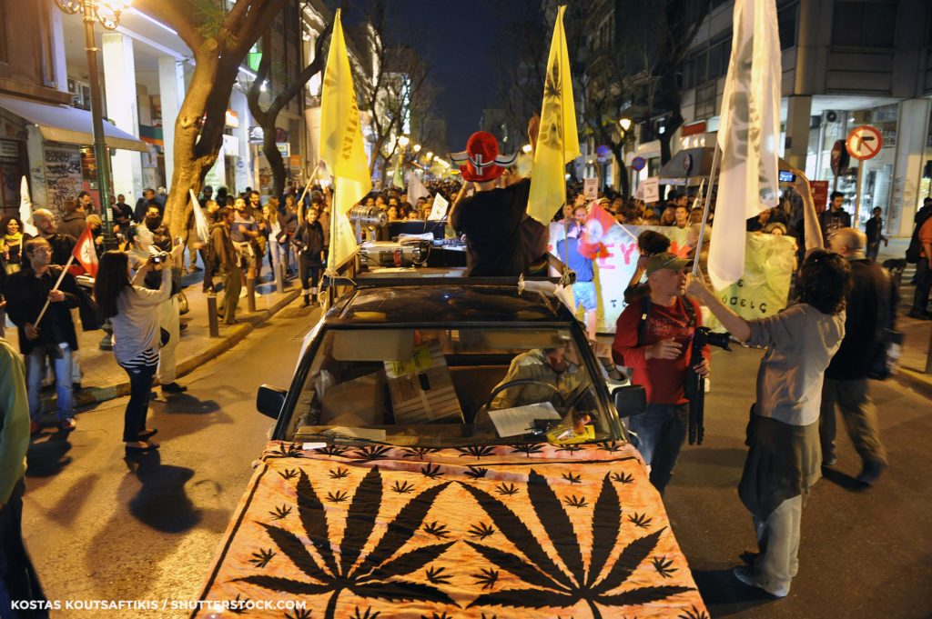 Un automóvil conduciendo a través de multitudes de personas con banderas y símbolos de cannabis en el capó.