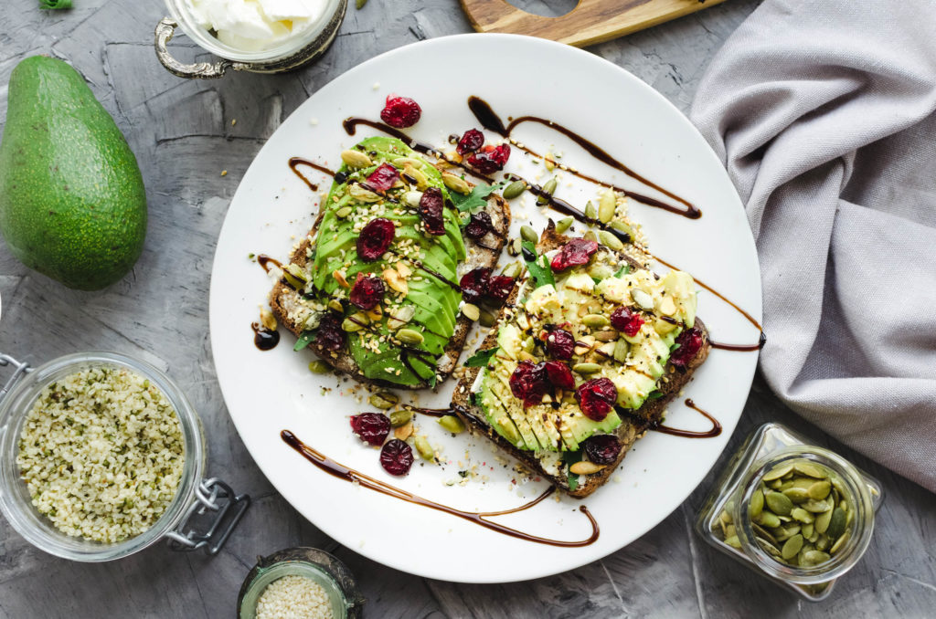 A white plate of avocado on toast topped with balsamic glaze, dried fruit and cannabis seeds