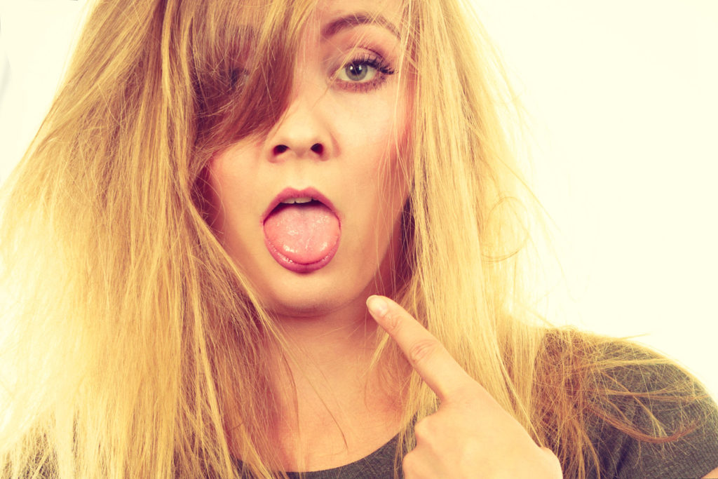 A blonde woman with her mouth open. She is pointing to her tongue