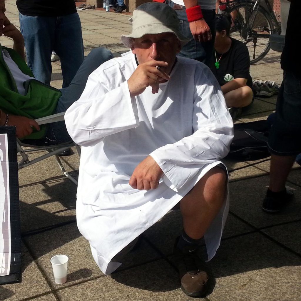 Ralf Herrmann in squat wearing a white robe smoking joint