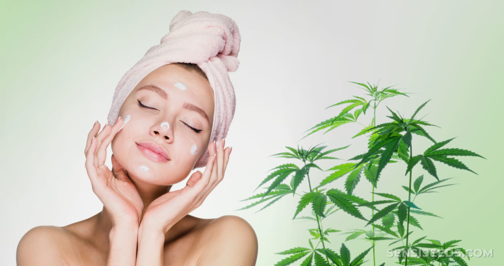 A woman with a towel on her head putting on skincare next to a cannabis plant