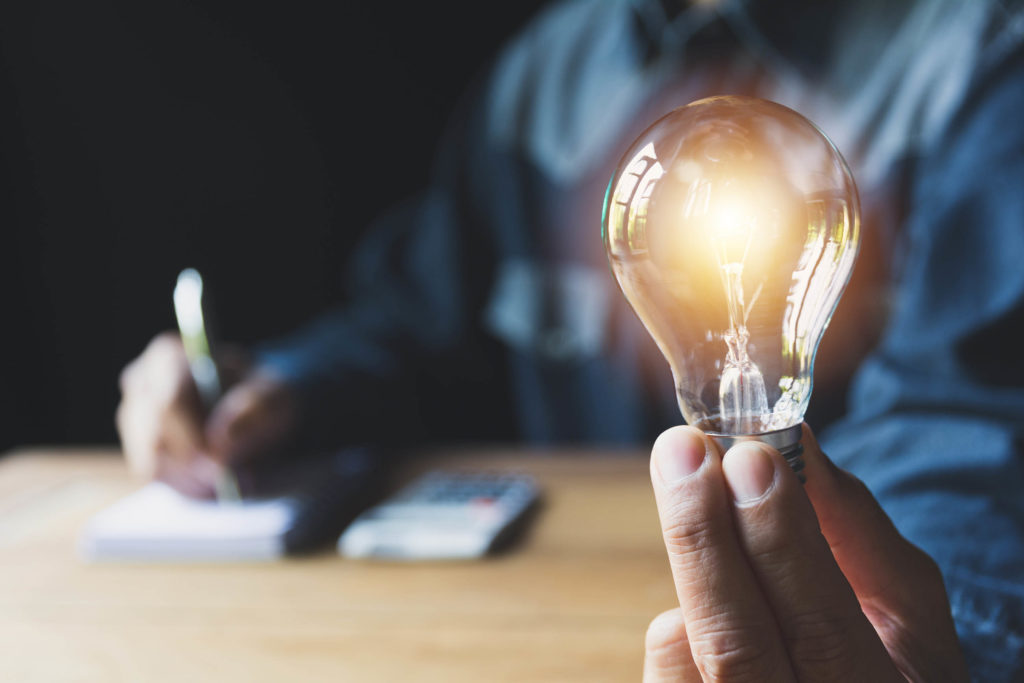 A person writing in a notepad with a calculator next to it and holding a lightbulb