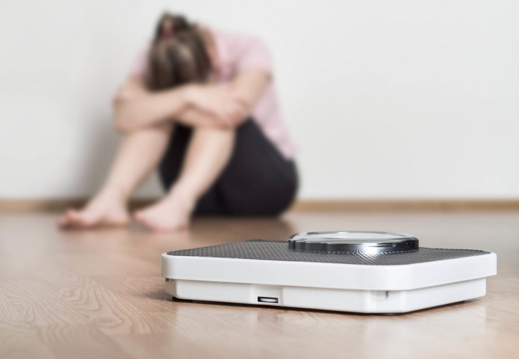 A weight scale and a woman hugging her knees in the background