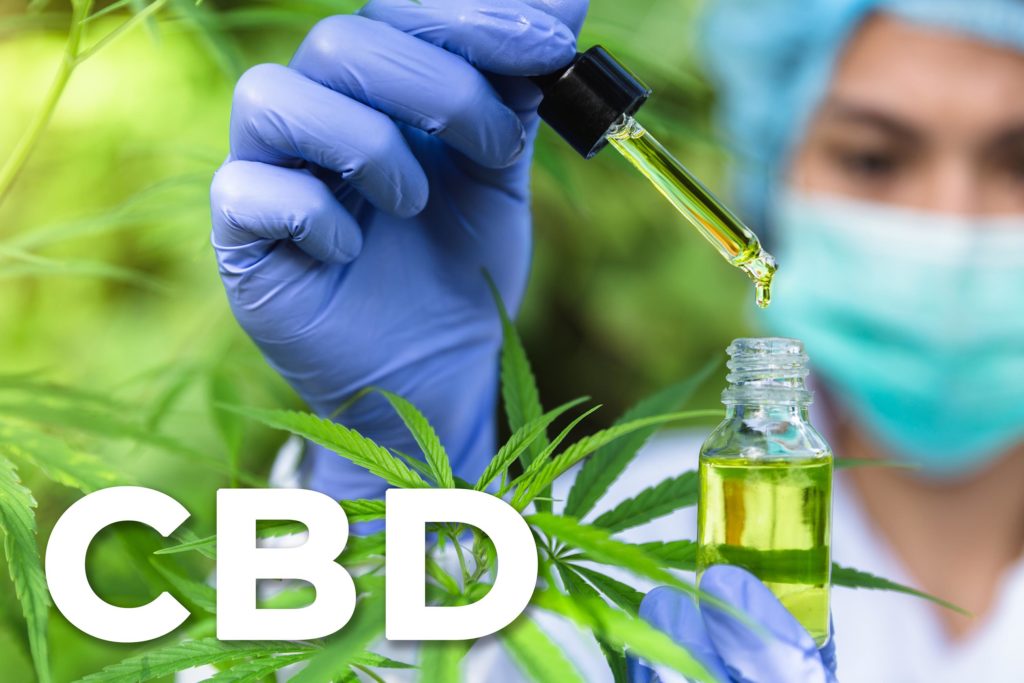 A person wearing a mask and gloves holding a pipette and bottle of CBD oil