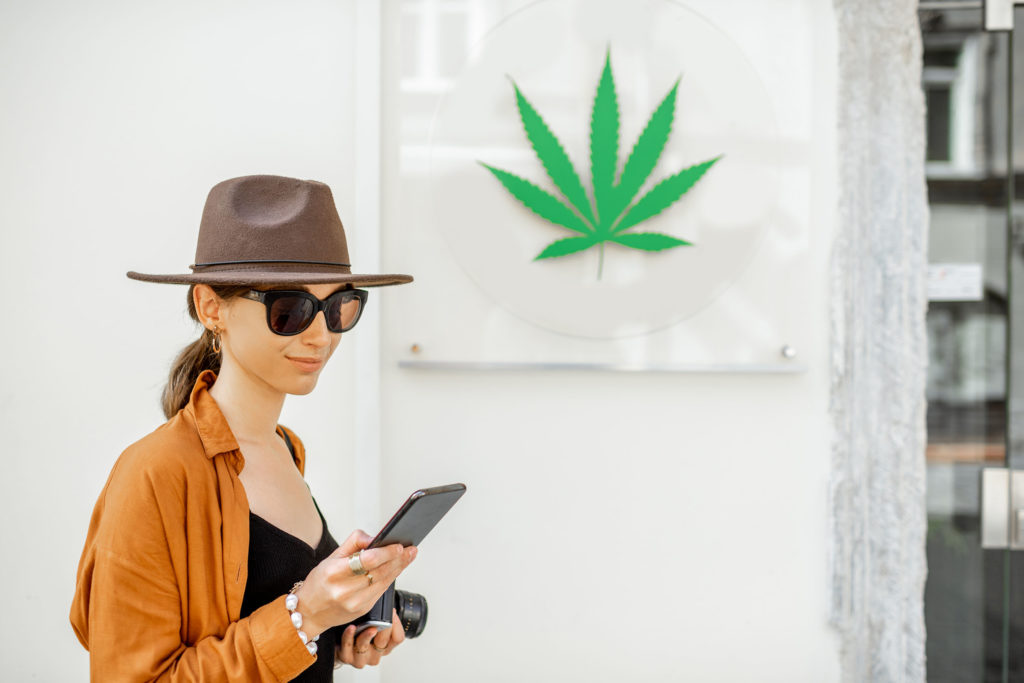 Woman with hat and sunglasses looking at mobile with cannabis leaf sign behind