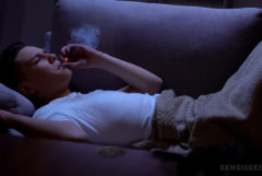 A man in white t-shirt, covered with beige blanket, lying on a sofa smoking a joint