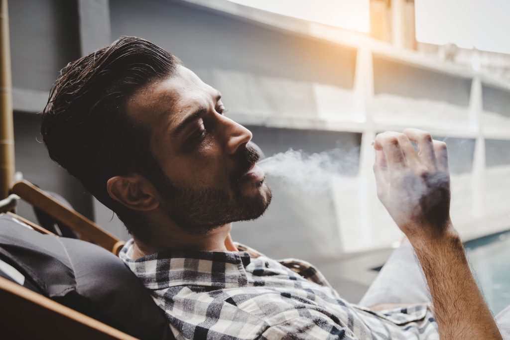 A man with brown hair and beard blowing a smoke from cannabis joint