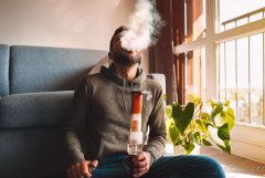 A man with a beard in a grey sweatshirt smoking a dub in a living room